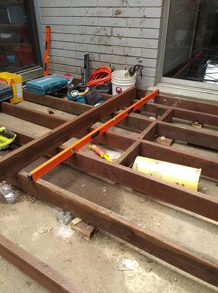 Orange level resting on wooden floor joists at a construction site beside a wall with tools and supplies.