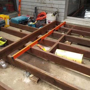 Wooden deck framing under construction with an orange level across beams beside a brick wall.