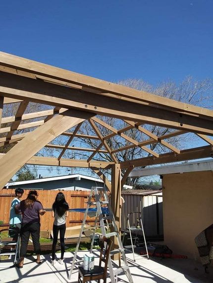 People on a patio under a wooden pergola frame, with a ladder and tools during construction.