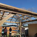 People building a wooden roof frame outdoors, with a ladder and blue sky overhead
