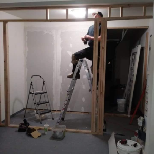 Person on a ladder working on drywall in an unfinished room with tools on the floor.