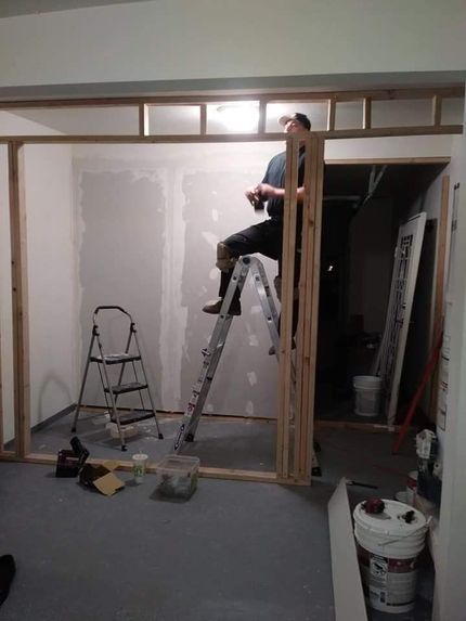 Person on a ladder working on unfinished interior wall with tools and buckets nearby.