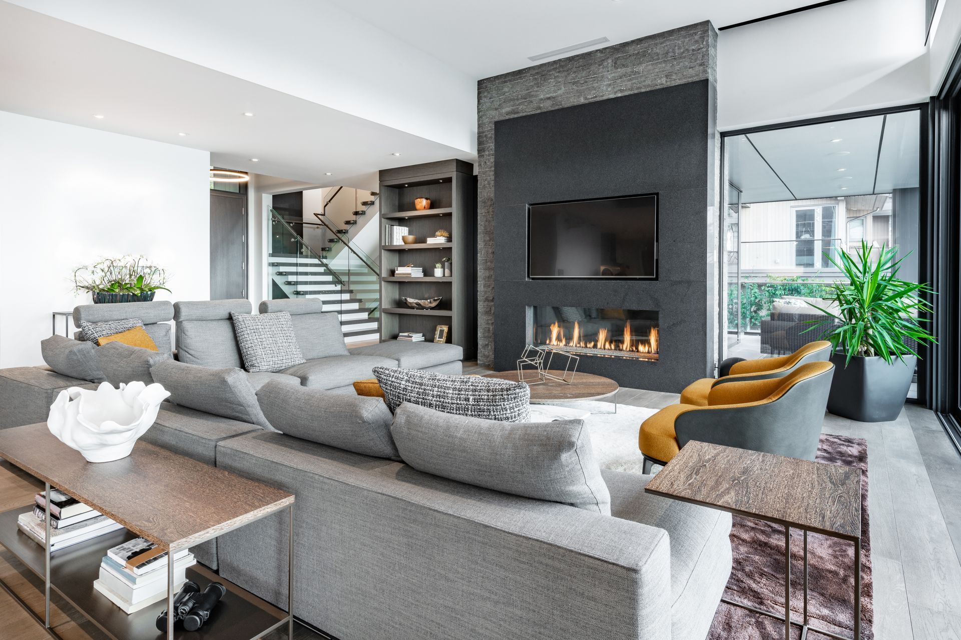 Modern living room with gray sectional sofa, black fireplace, and large windows overlooking greenery