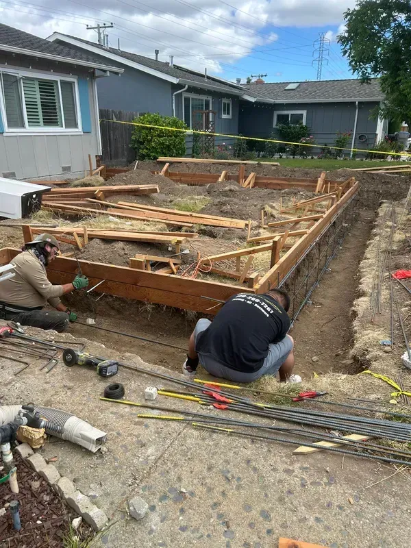 Workers laying a foundation with rebar and wooden forms beside a house in a gravel yard