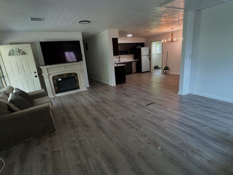 Open living room with fireplace, TV, gray sofa, and kitchen area, featuring light wood flooring and white walls.