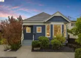 Blue house with gray roof, front porch, and shrubs at dusk under a pink sky