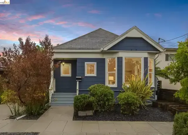 Blue house with gray roof, front porch, and shrubs at dusk under a pink sky