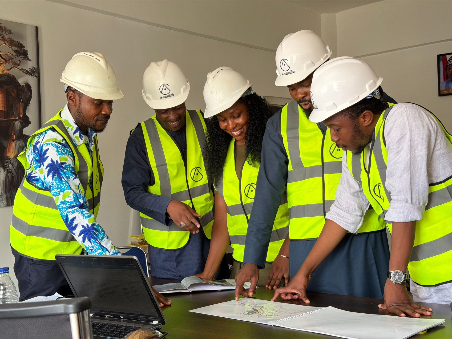 Five workers in hard hats and safety vests review plans around a laptop in a meeting room