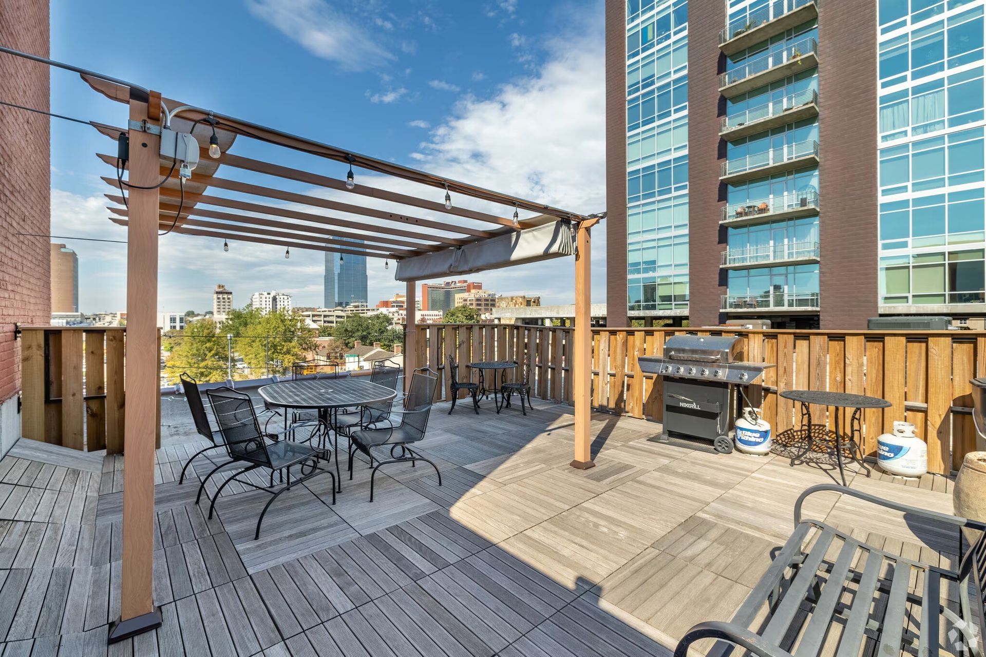 Rooftop patio with seating, grill, and pergola, overlooking city buildings on a sunny day.