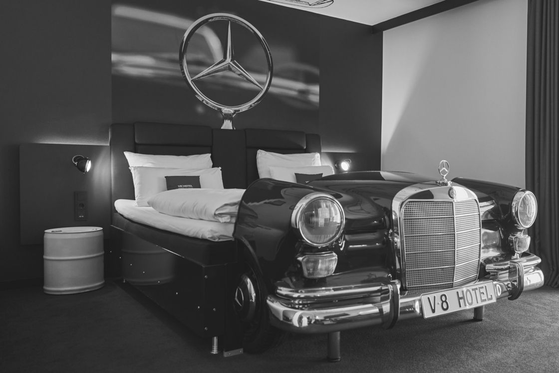 Ein Oldtimer-Mercedes parkt in einem Hotelzimmer unter einem Mercedes-Logo-Wandschild.