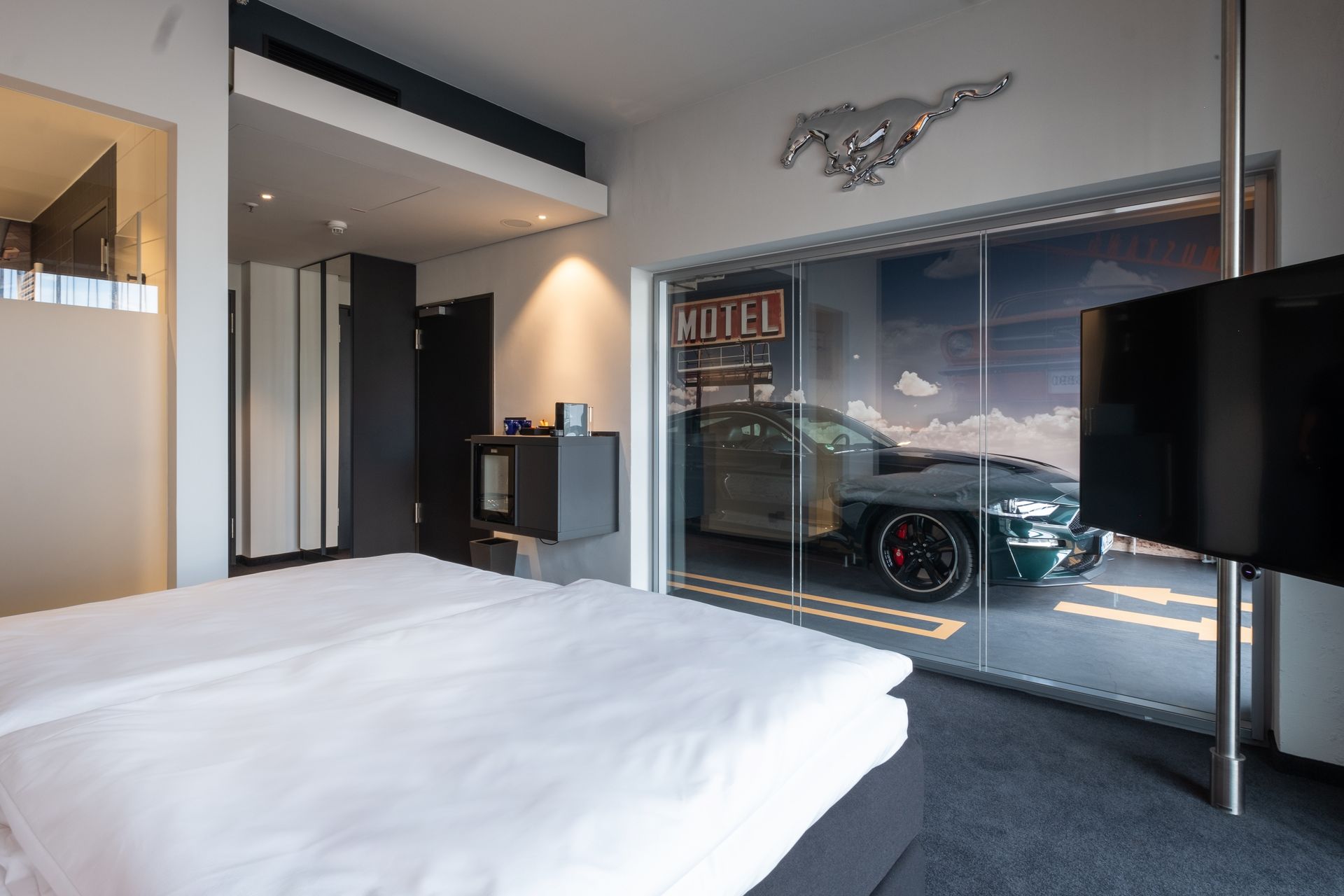 Modernes Hotelzimmer mit Bett, Fernseher und einer Glaswand mit Blick auf eine Ausstellung geparkter Autos.