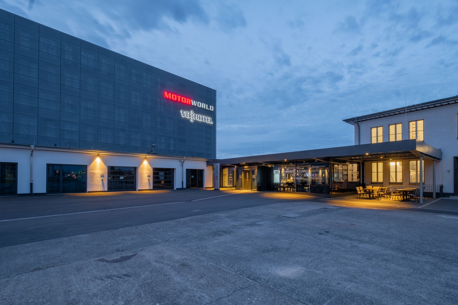 Abenddämmerung: Außenansicht eines modernen Hotels mit beleuchteten Bögen, Kiesparkplatz und einem leuchtenden Eingangsplatz.
