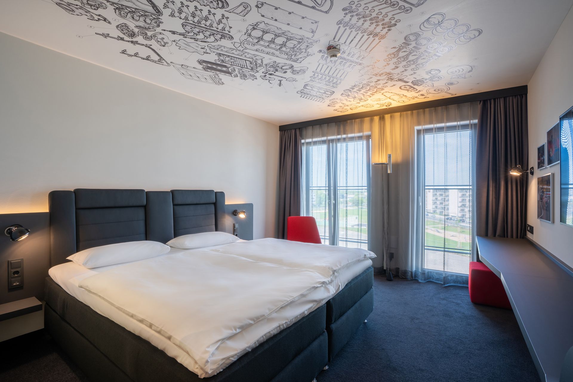 Modernes Hotelzimmer mit großem Bett, gemusterter Decke, rotem Sessel und Balkontüren mit Vorhängen.
