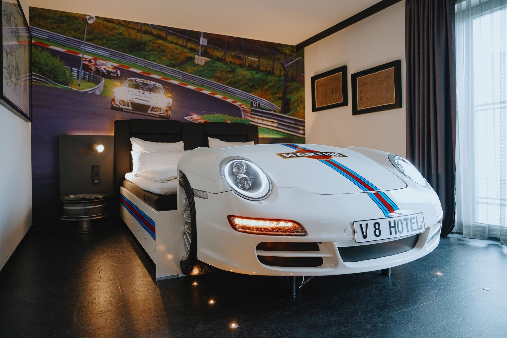 Ein weißer Sportwagen, der in einem modernen Hotelzimmer als Bett gestaltet ist, mit Rennsport-Wandbild und schwarzen Vorhängen.