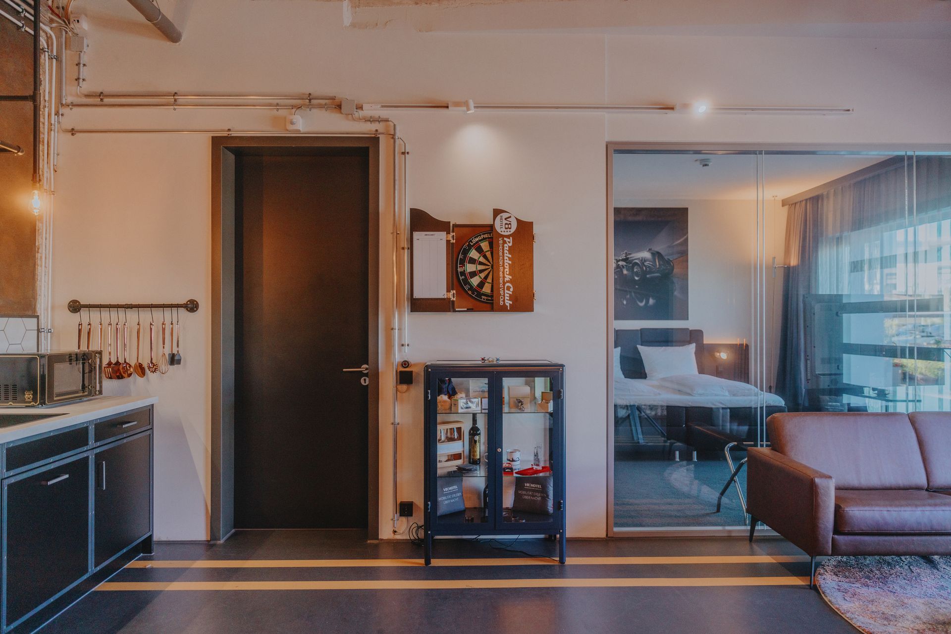 Modernes Wohnzimmer mit Küche, dunkler Tür, Wandbildern und einem großen Spiegel, der ein Sofa und die Fenster reflektiert