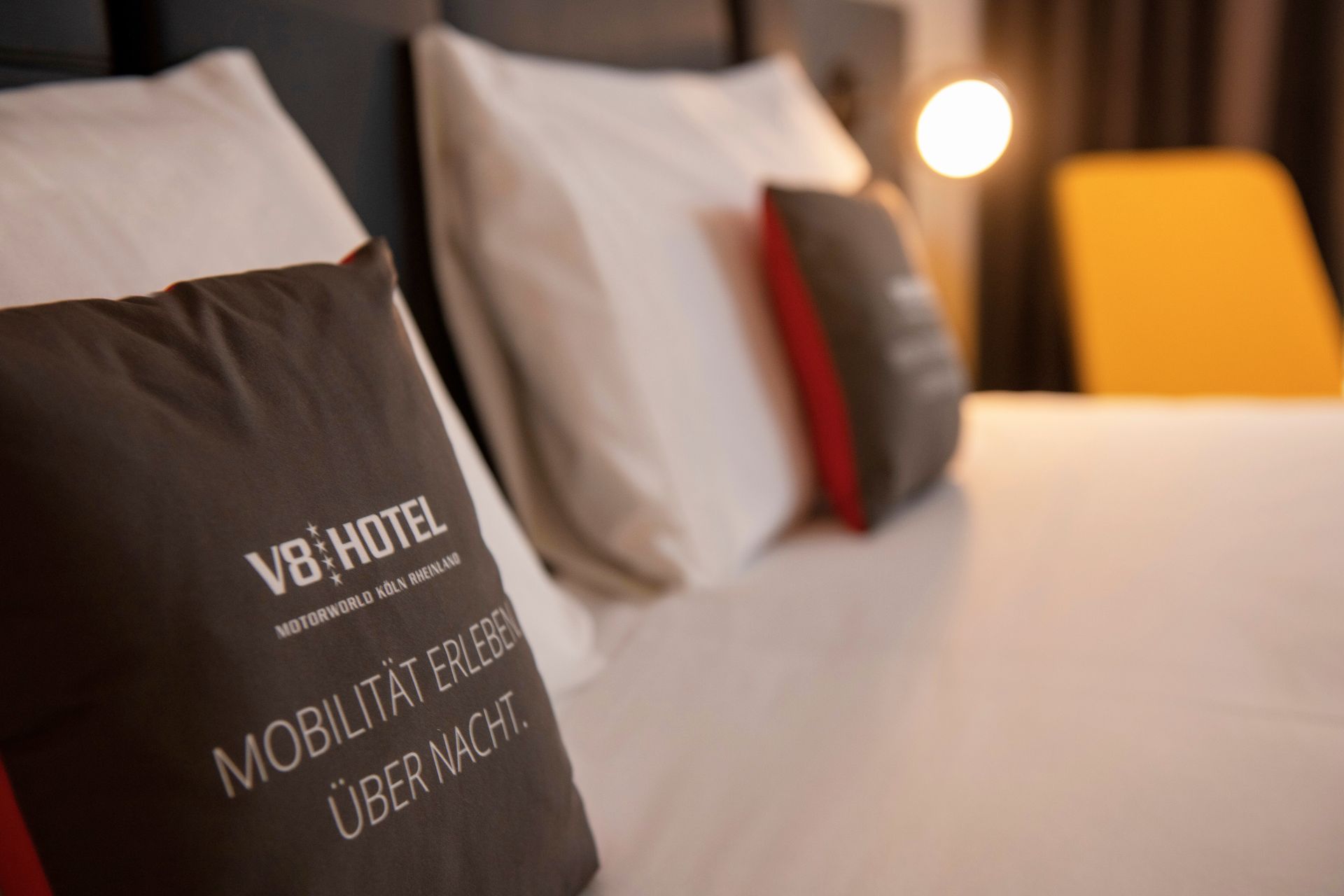 Hotelbett mit dunkelgrauen Kissen, weißen Laken und einer warmen Nachttischlampe in einem sanft beleuchteten Zimmer