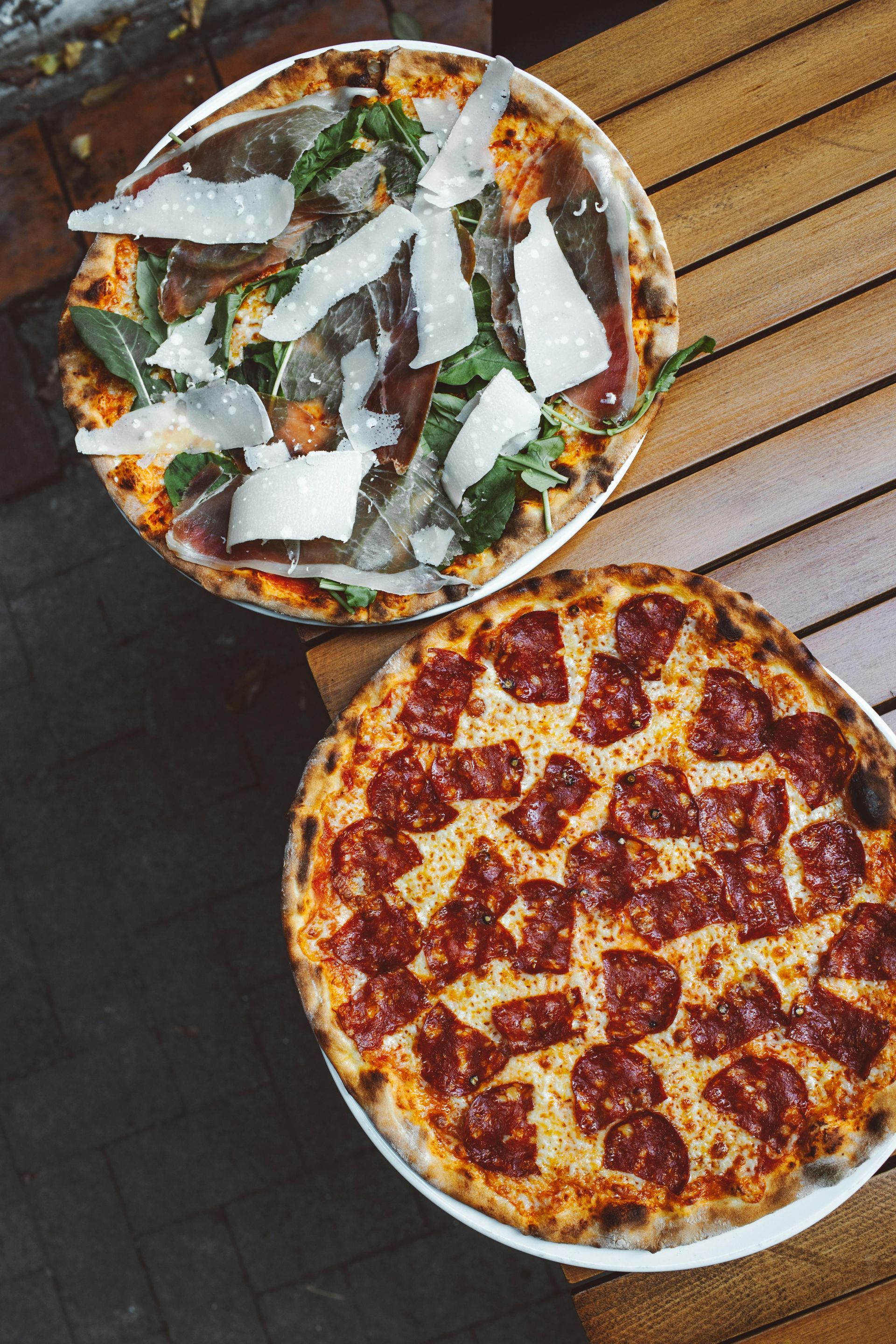 Zwei Pizzen auf einem Holztisch: eine mit Rucola und gehobeltem Käse, eine mit Peperoni.