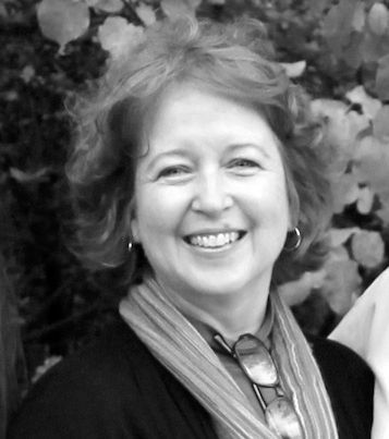 Woman smiling outdoors, wearing glasses on a scarf, with trees in the background.