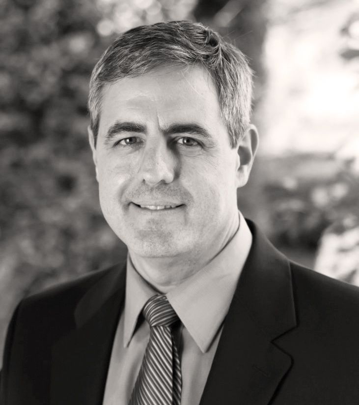 Man in suit smiles at the camera, outdoors with blurred greenery background. Todd Hendrix