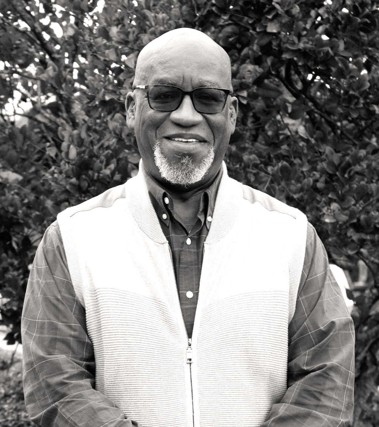 Man wearing glasses, collared shirt, and vest, smiling in front of foliage. Bernard Anderson