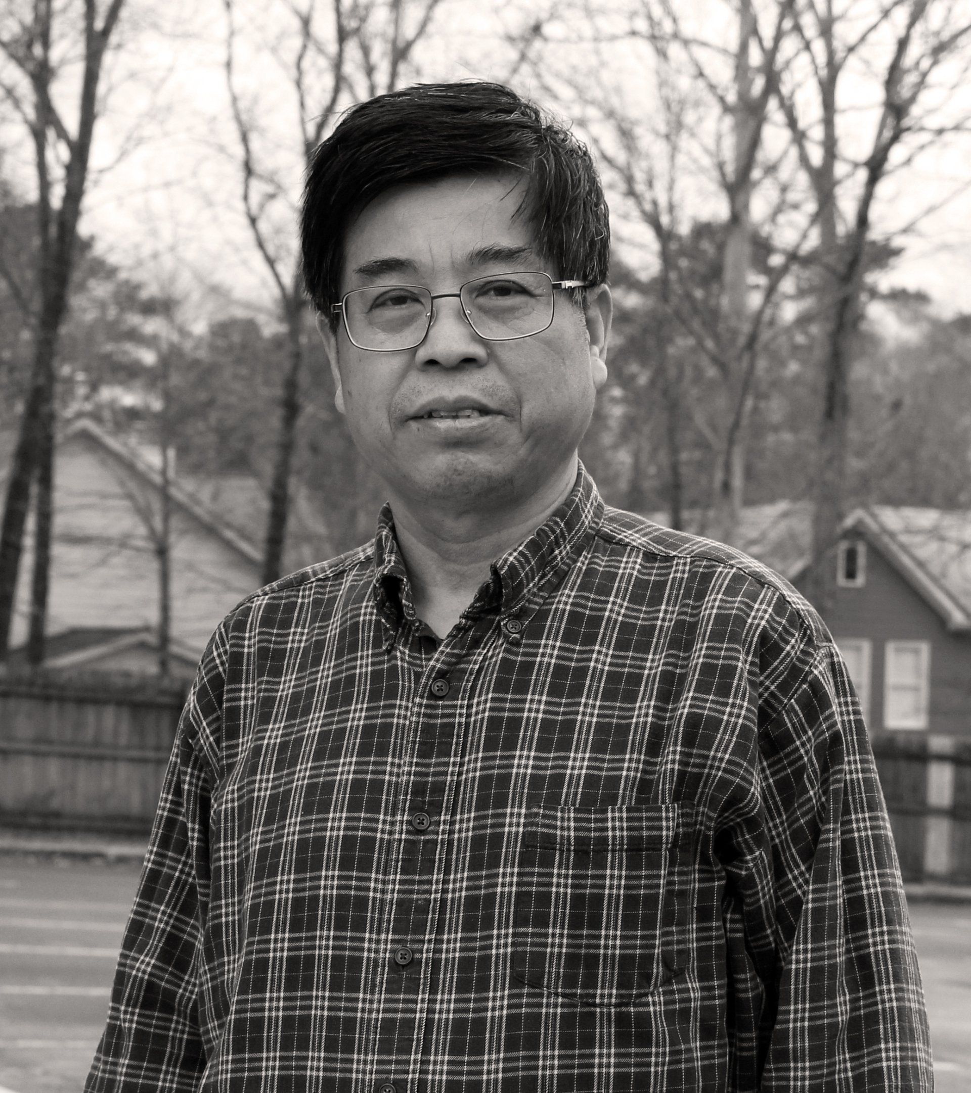 Man in plaid shirt and glasses standing outdoors, blurred houses in background. Faun