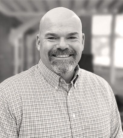 Bald man with a beard smiling, wearing a button-down shirt. Blurred indoor background. Andrew Keasling
