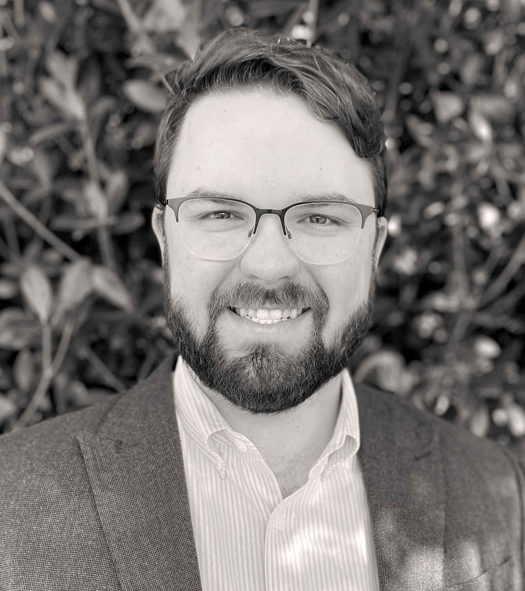 Man with glasses and beard smiles, wearing a blazer, against a leafy backdrop. Braden Benson