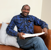 Man in blue plaid shirt smiles while holding a book, sitting on brown leather couch.