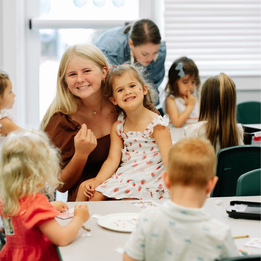 A smiling blonde child and another child sit at a table in a classroom with other children and an adult.