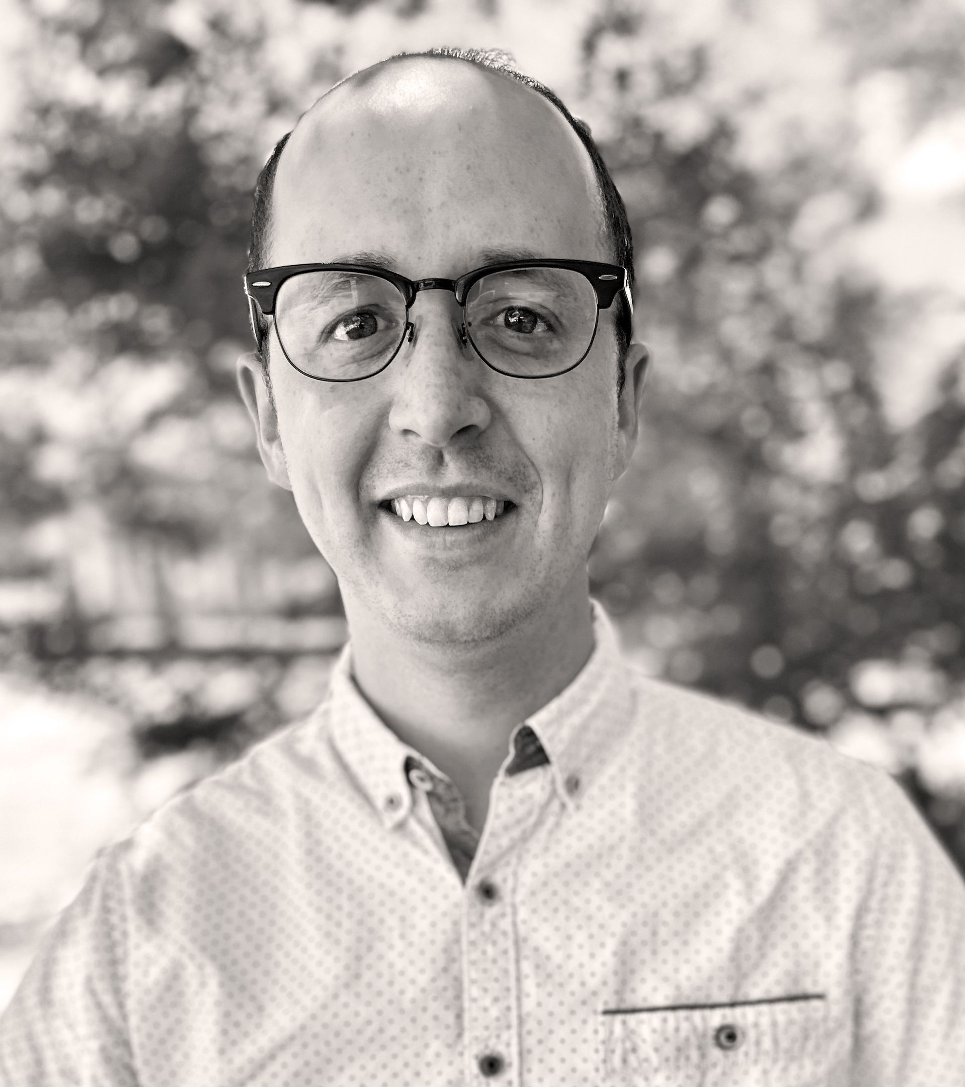 Man with glasses smiles at the camera, wearing a button-down shirt outdoors. Erik Lundeen 