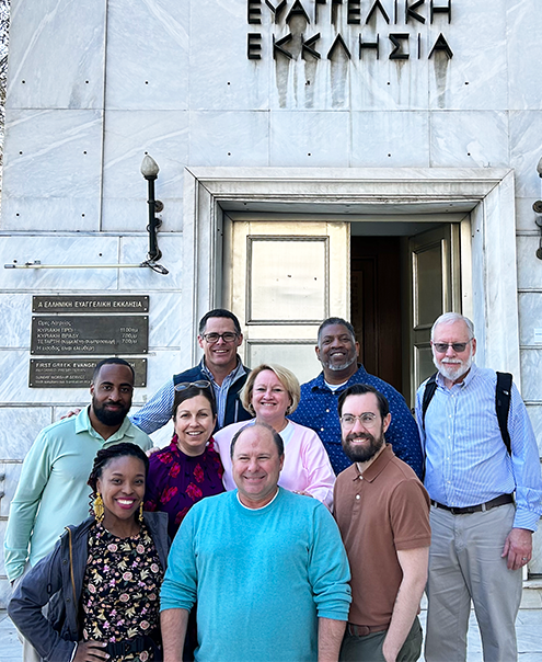 Group of people standing in front of a building with Greek text. People smiling, wearing various clothing.
