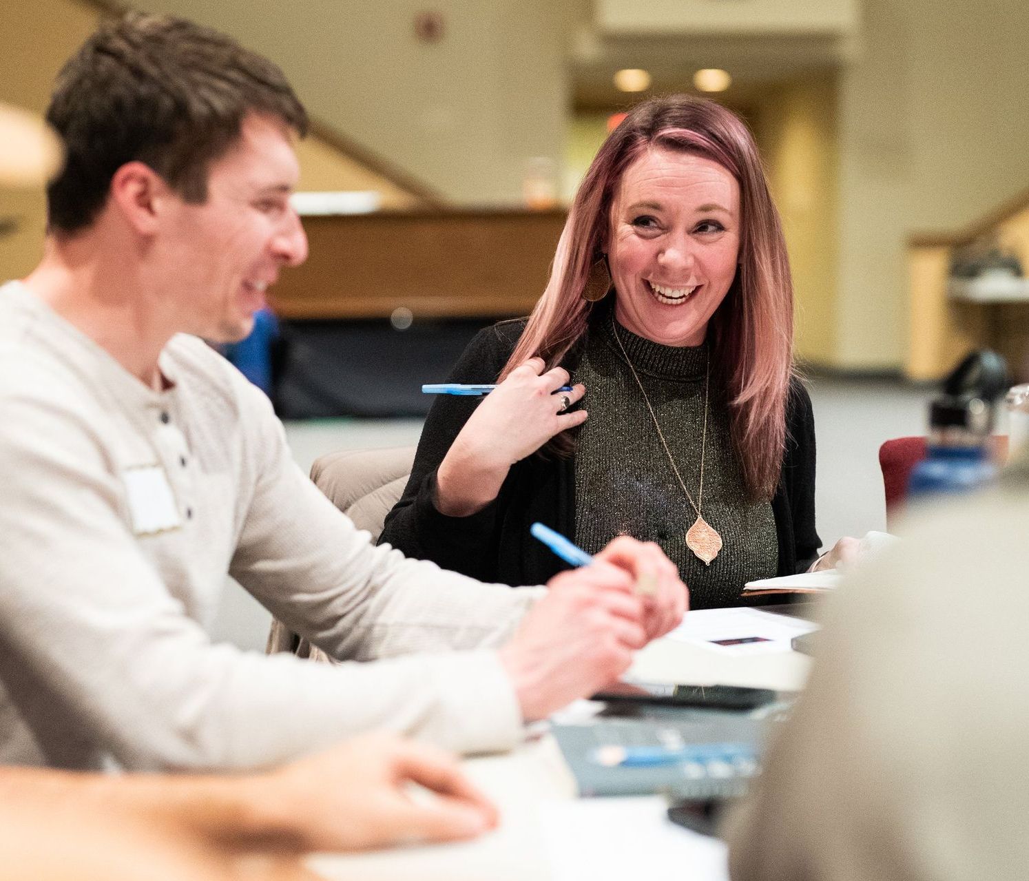 Two people laughing while sitting at a table, possibly in a meeting; woman with pink hair, man in a light gray shirt.