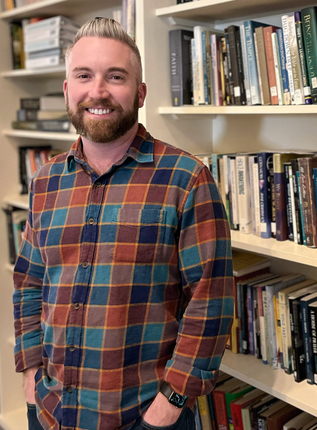 Man with a beard, wearing a plaid shirt, smiles in front of a bookshelf filled with books.