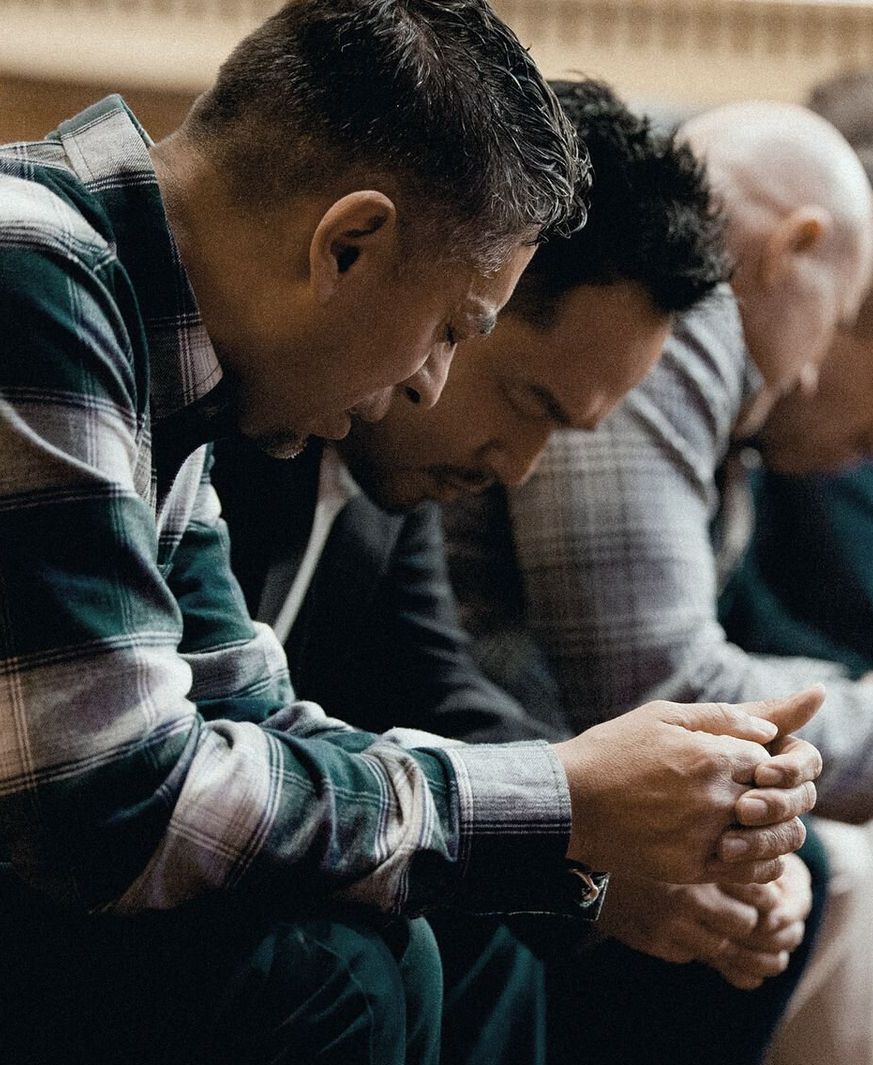 Men bowing their heads in prayer, hands clasped, indoors.