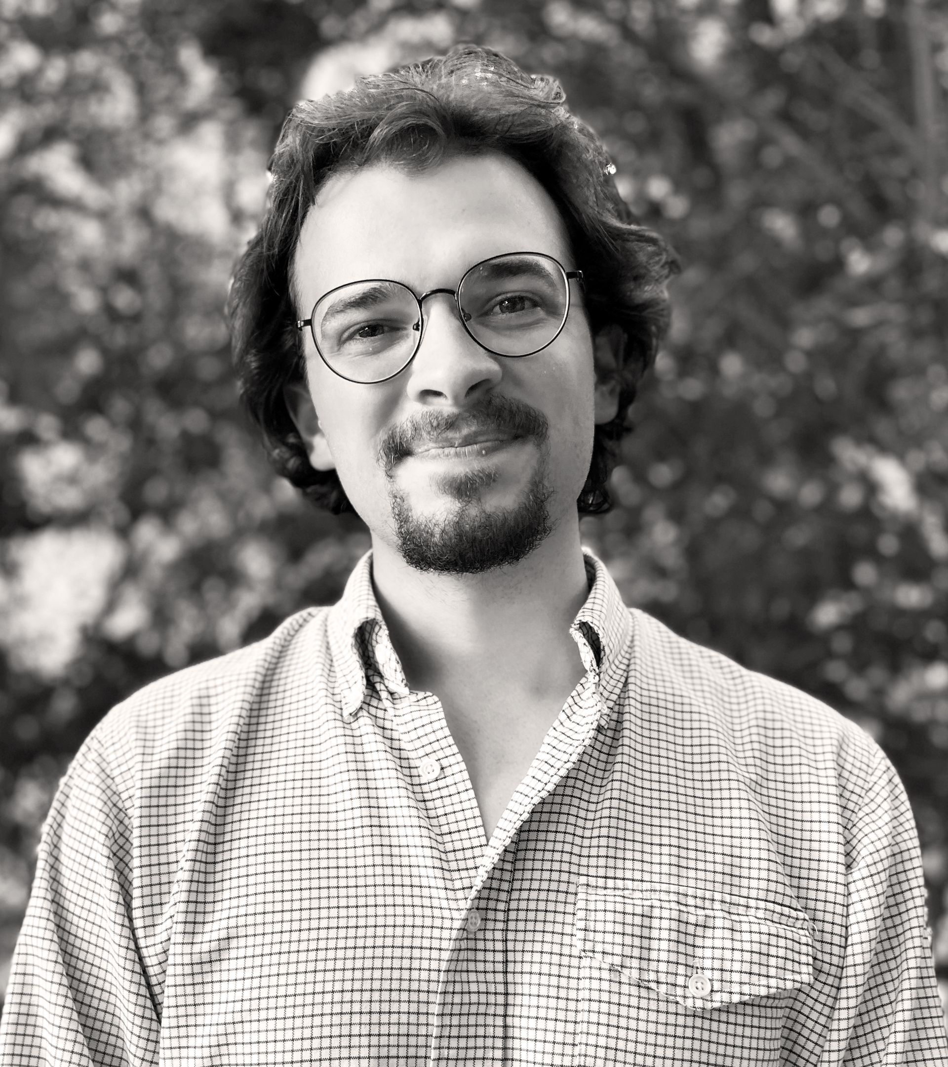 Man with glasses and beard, smiling. Wearing button-up shirt, outdoors with blurred greenery. Sawyer McGonagill