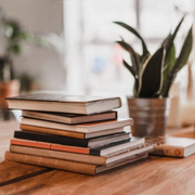 Pile of books on a wooden table, with a potted plant in the background. Natural light.