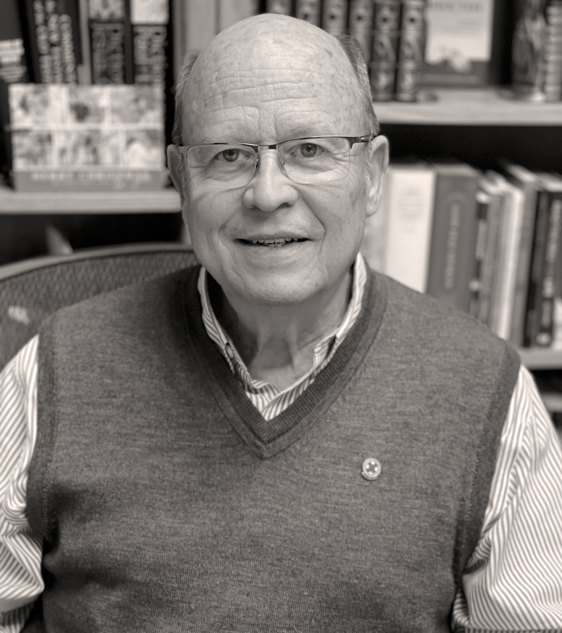 Man with glasses and a sweater vest smiles, bookshelves in the background. Tom McClendon