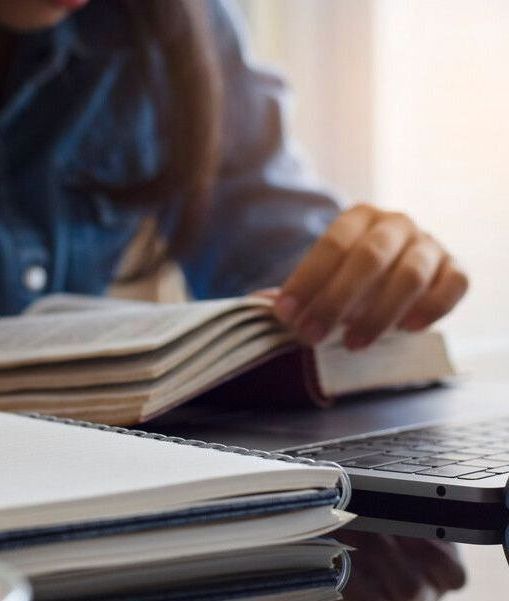 Person reading a book at a desk with a laptop and notebooks.
