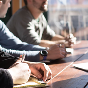 Three people at a table; one writes in a notebook, others look on. Sunny day.