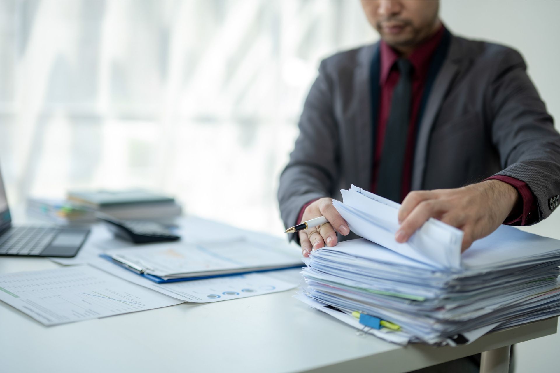 Person in suit sorting through a stack of papers on a desk with a laptop and documents.