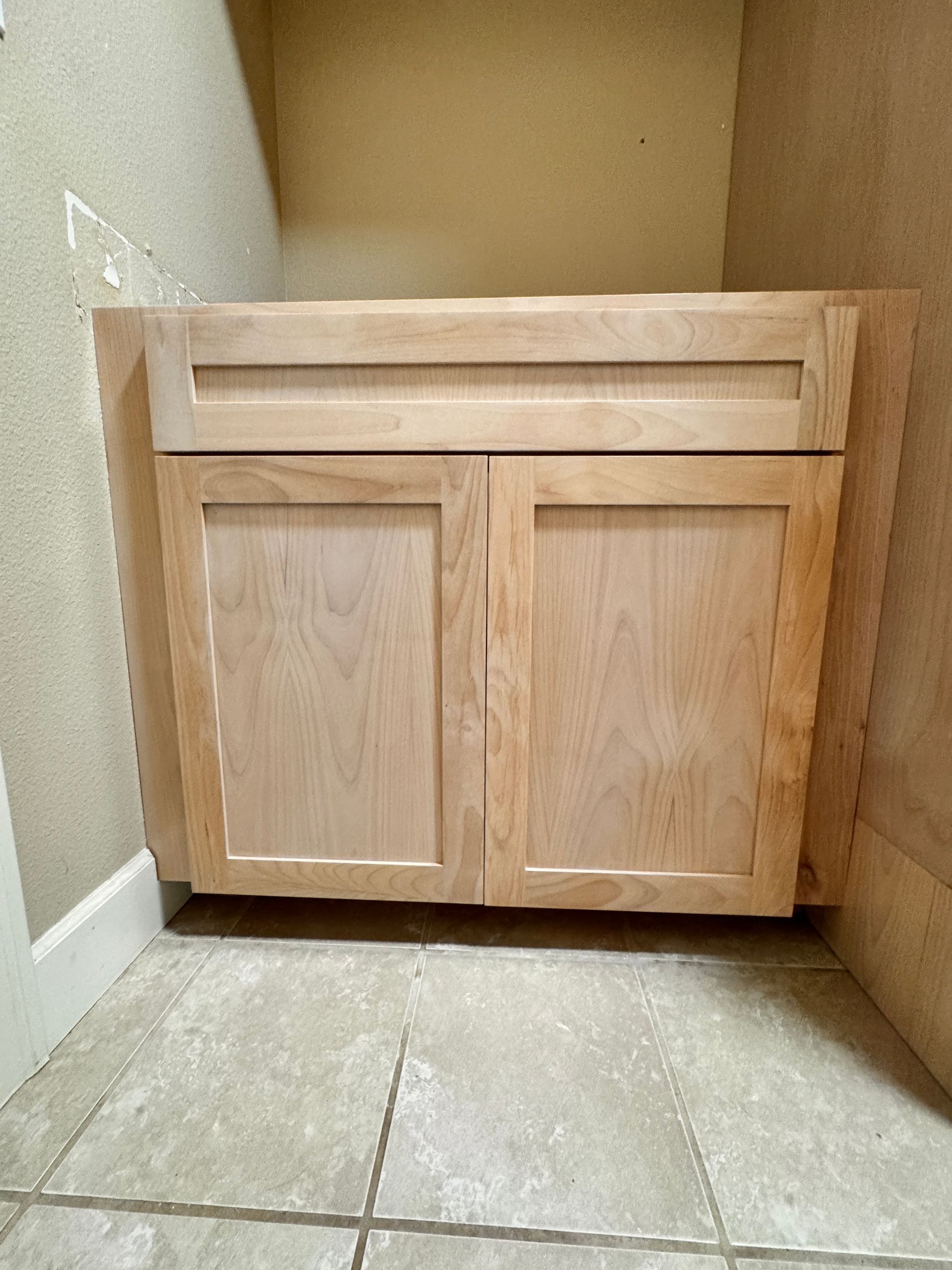 Wooden bathroom vanity cabinet with two doors and a drawer, placed in a tiled corner.