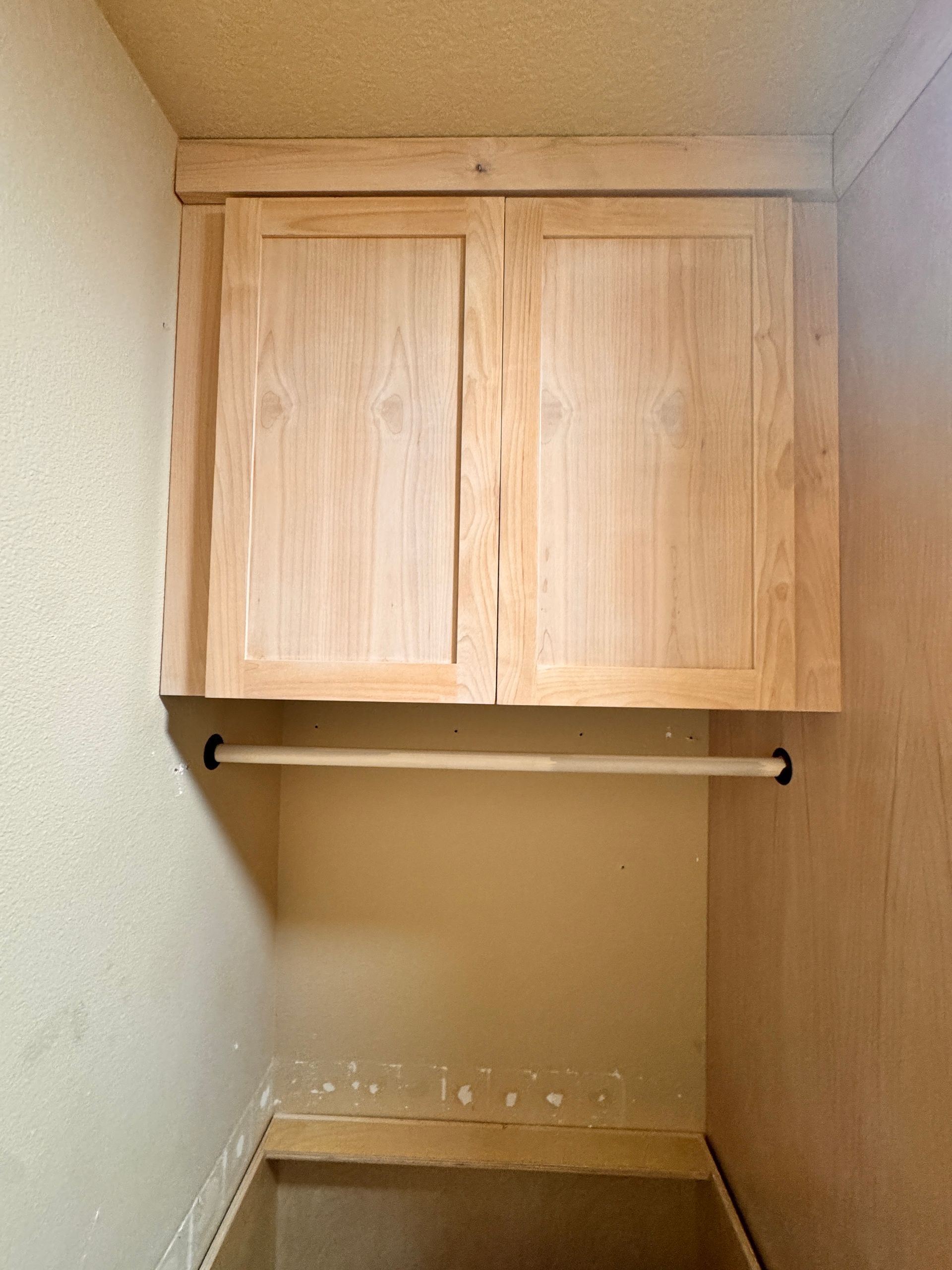 Small closet with wooden cabinet doors, a hanging rod, and empty beige walls.