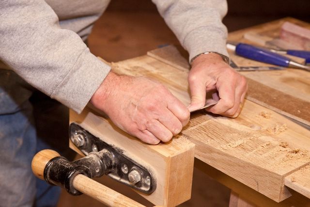 Hands planing a wooden board on a workbench in a woodworking shop.