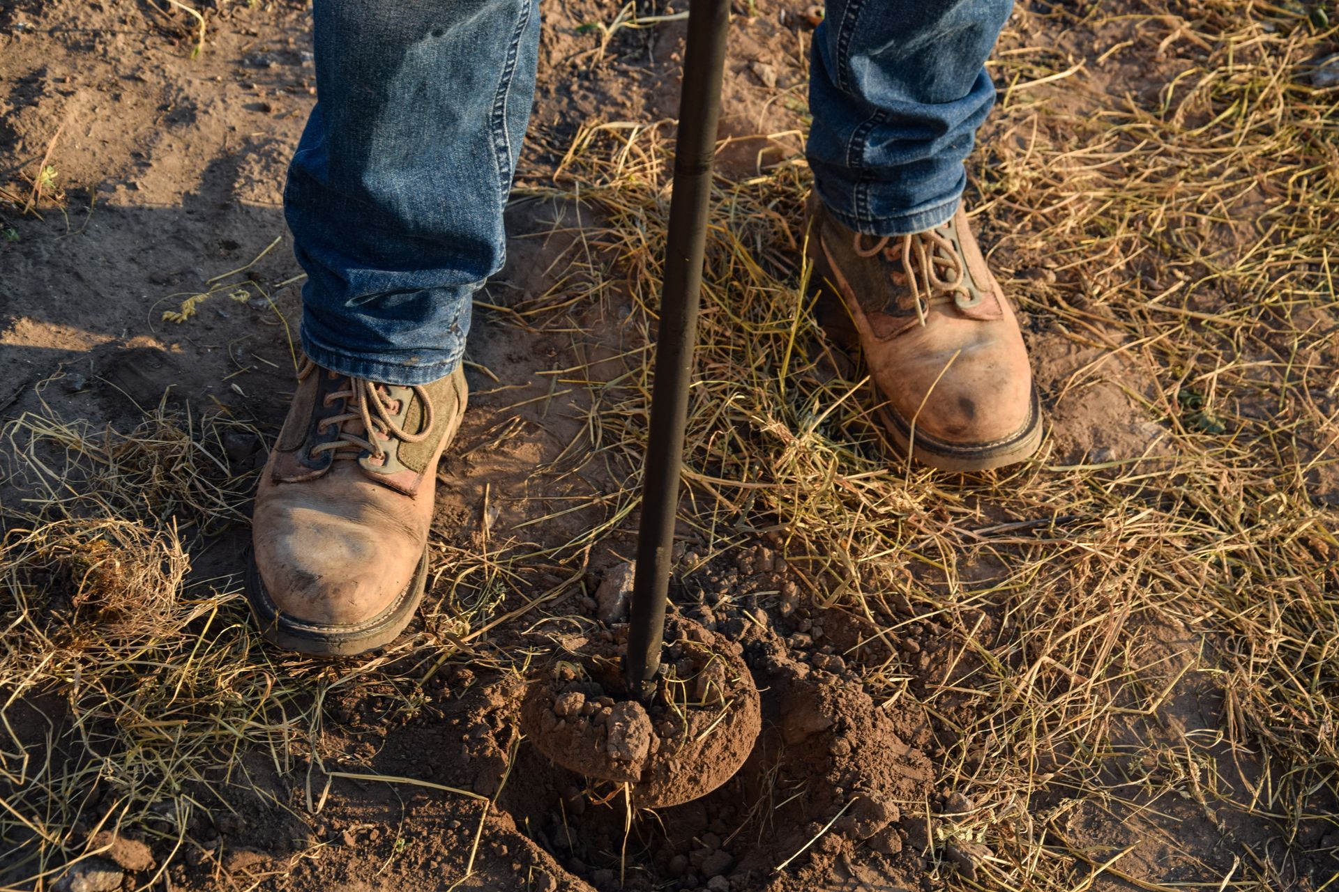 Person in jeans and work boots standing by a shovel in dry, grassy soil.