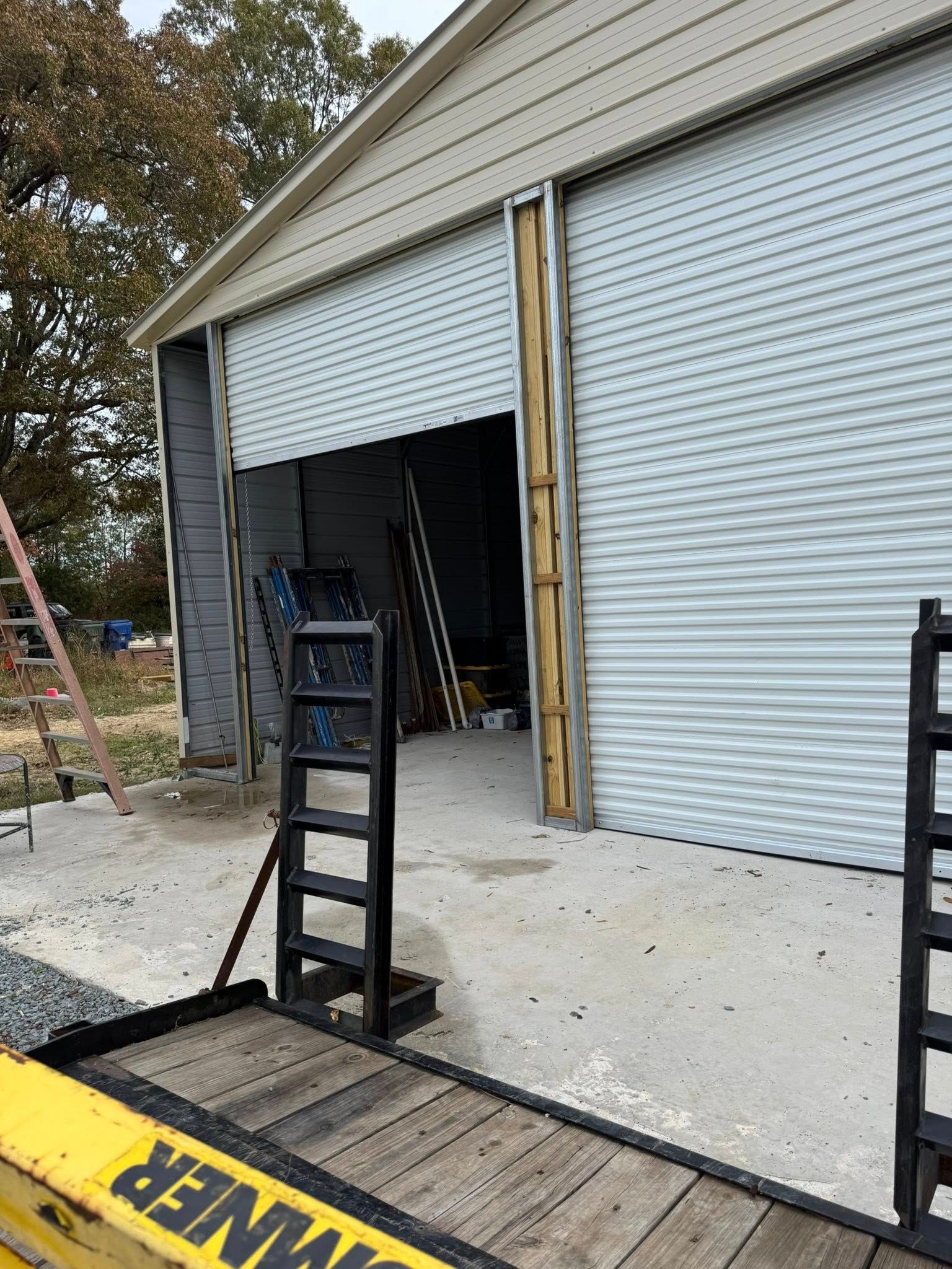 Partially open garage with white roll-up door, ladders, and a loading trailer in front