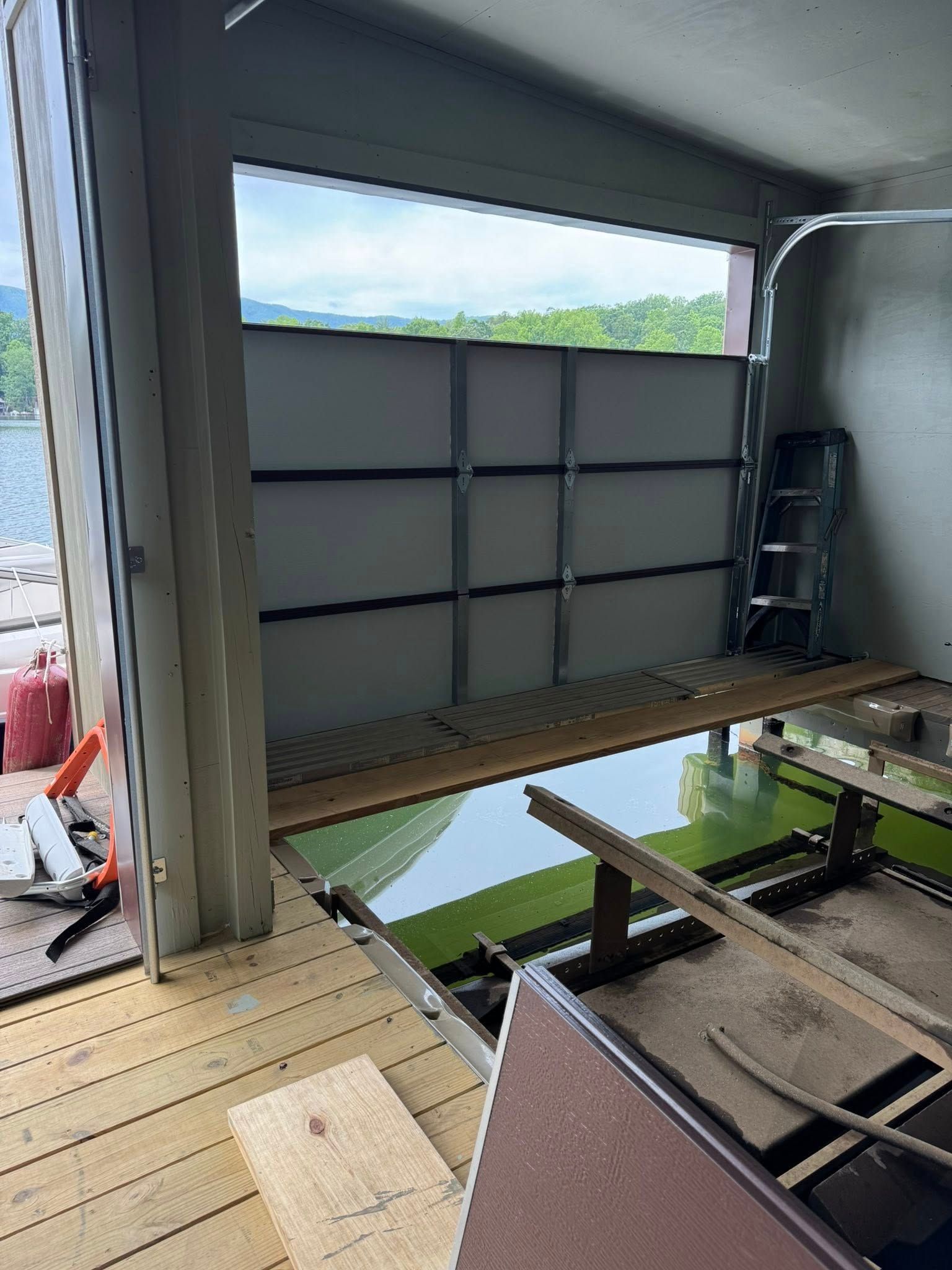 Interior of a partially open garage with a large horizontal door, wooden deck floor, and a leaning ladder.