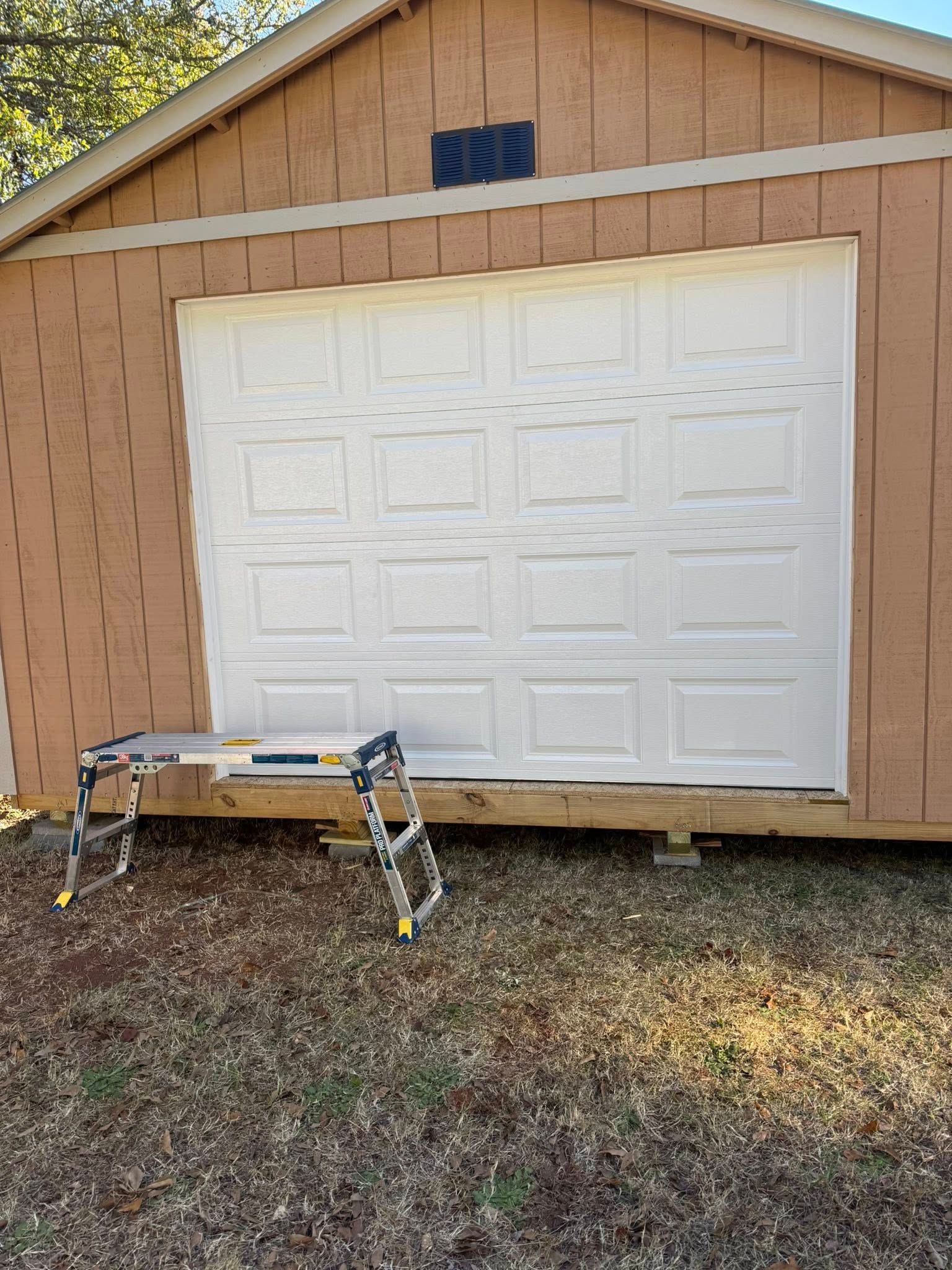 White garage door on tan building with a small work platform in front and gravel ground.