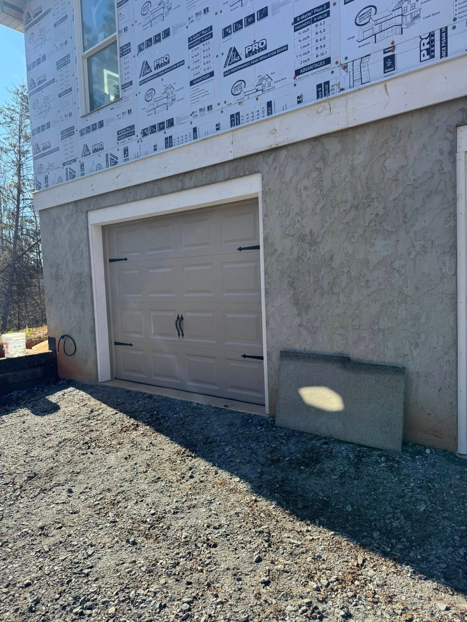 Concrete garage door on a house exterior with exposed sheathing, gravel ground, and a utility box beside it
