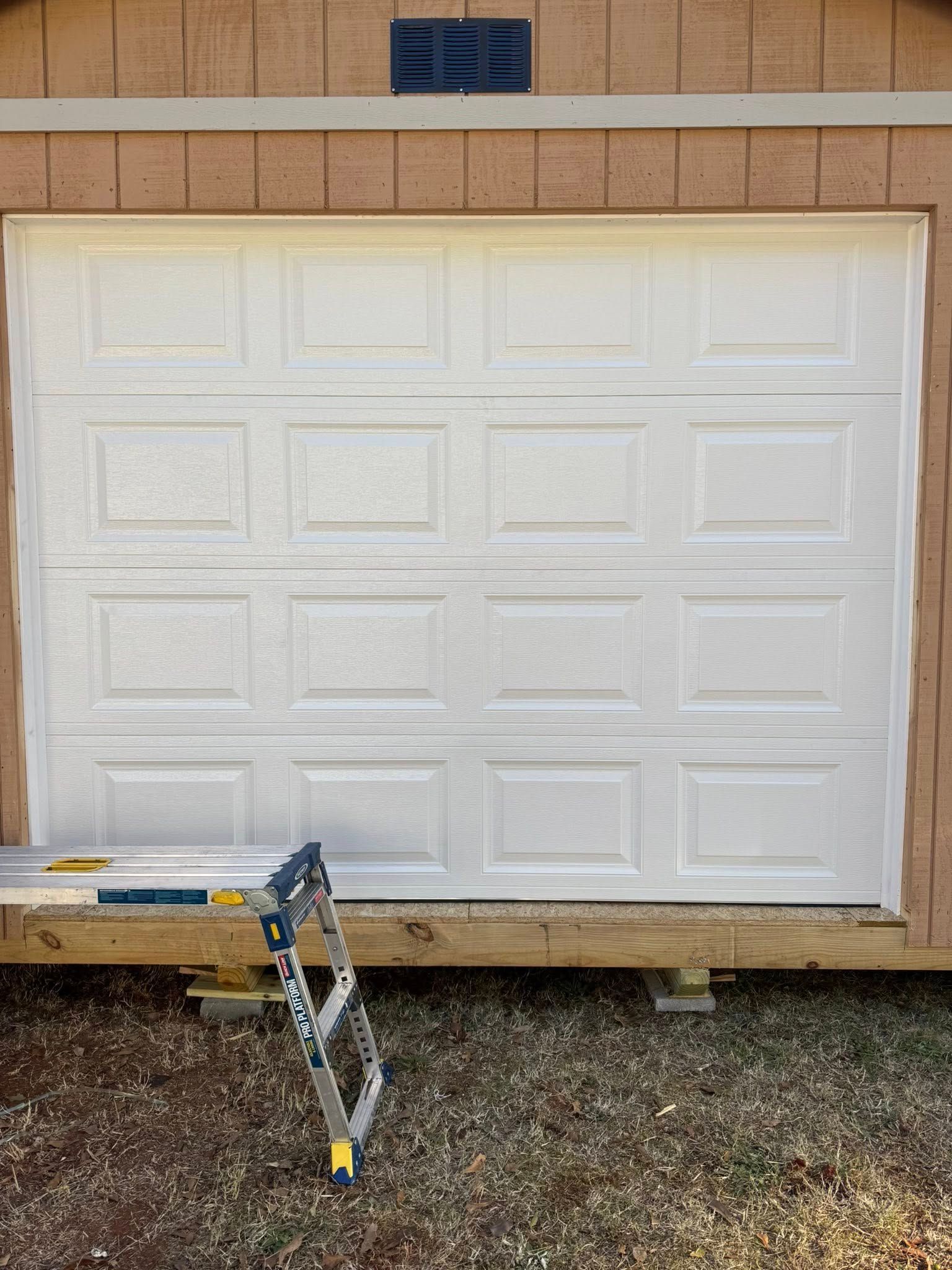 White garage door on a beige building, with a ladder and scaffold in front on gravel.