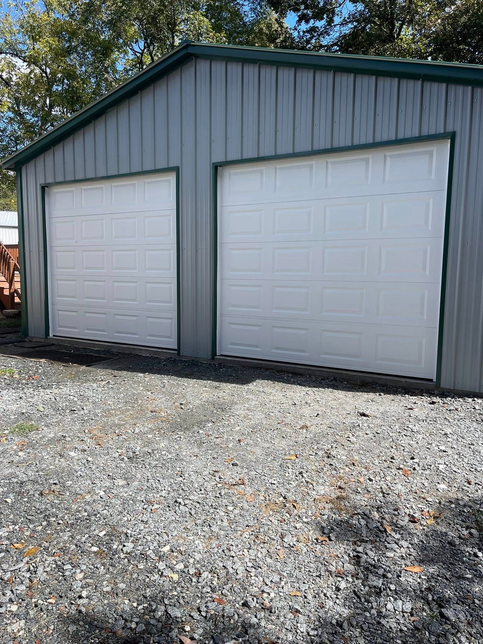 Gray metal two-car garage with white doors on a gravel driveway under trees